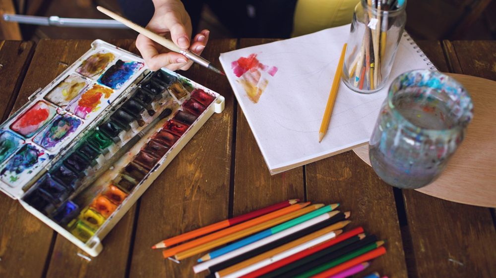 Watercolor paint box, colored pencils and a sketchbook on a wooden table
