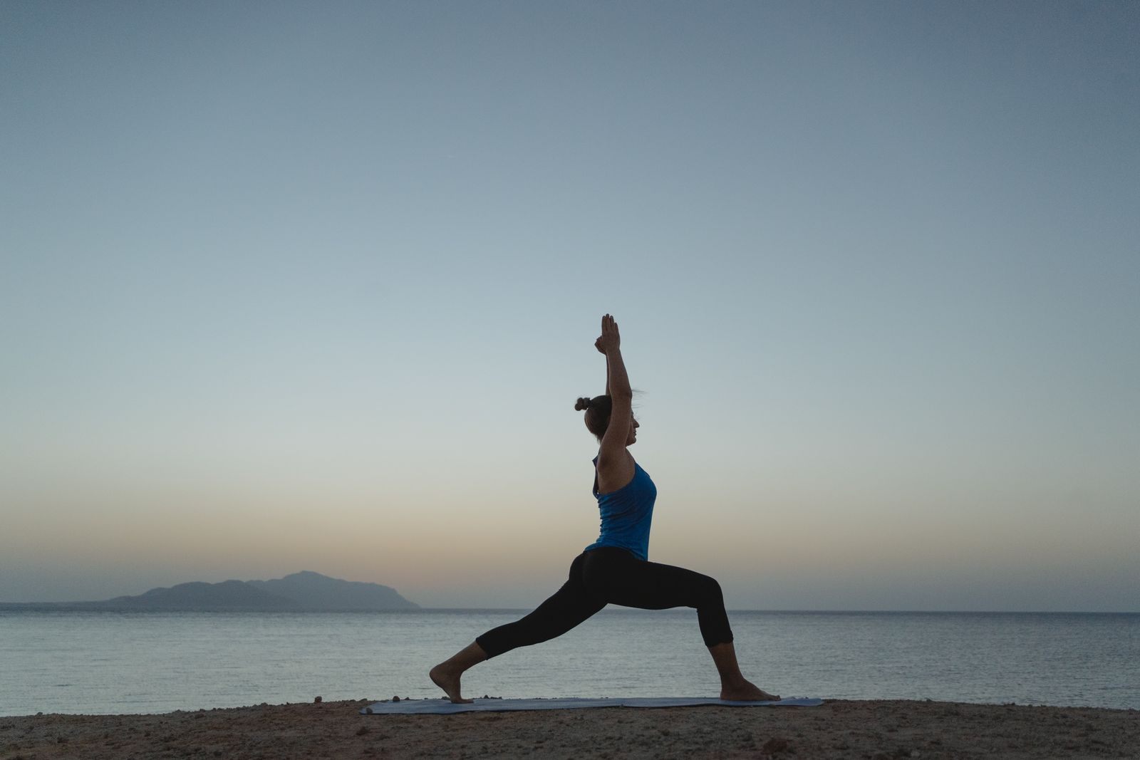 Woman in a warrior yoga pose by the sea at dawn, the setting of a yoga cruise morning class